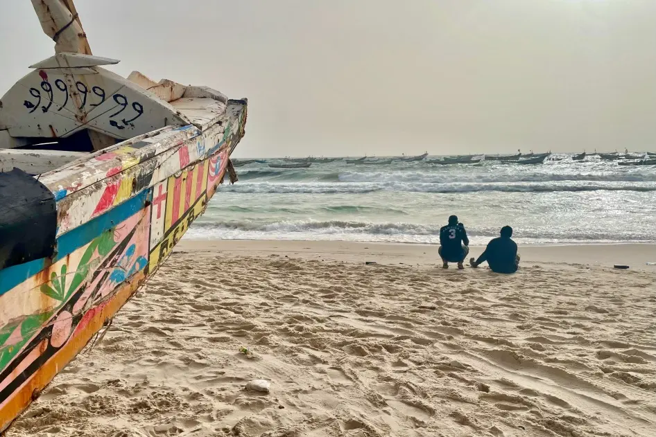Dos hombres cerca de una embarcación de madera conocida como piragua, utilizada tradicionalmente para la pesca en Mauritania y África Occidental, en una playa de Nuakchot, Mauritania, el 28 de junio de 2022. Las piraguas han sido utilizadas con frecuencia por personas migrantes que intentan cruzar el océano Atlántico para llegar a las islas Canarias, en España. © 2022 Lauren Seibert/Human Rights Watch